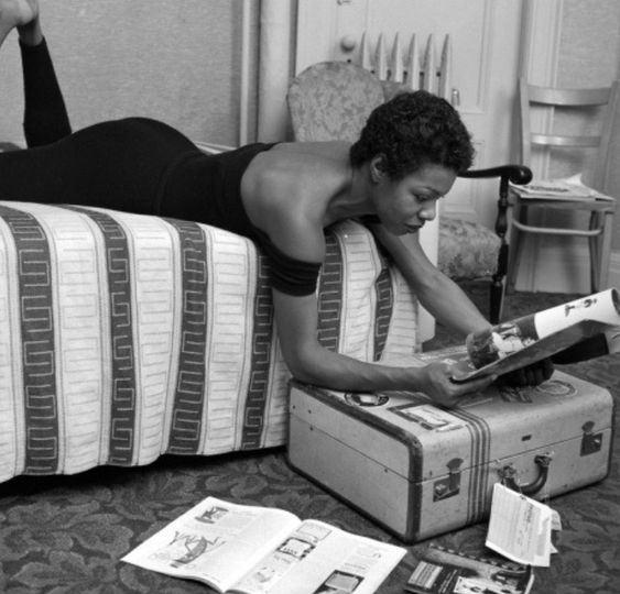 Maya Angelou in a off the shoulder black dress reading a magazine indoors