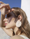 Model blocks sun from her face in a delicate manner as she wears large cream oval cotton earrings  calling attention to the braided technique