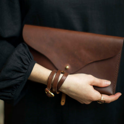 Woman holds a chestnut colored leather clutch against her black dress while wearing a matching leather bracelet