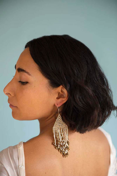 Short haired brunette model is slightly turned with her eyes looking down as her hand beaded earring gently rests on her exposed back. The earrings are hand beaded from gold and white crystals for Huichol Center by Wixáika artisans
