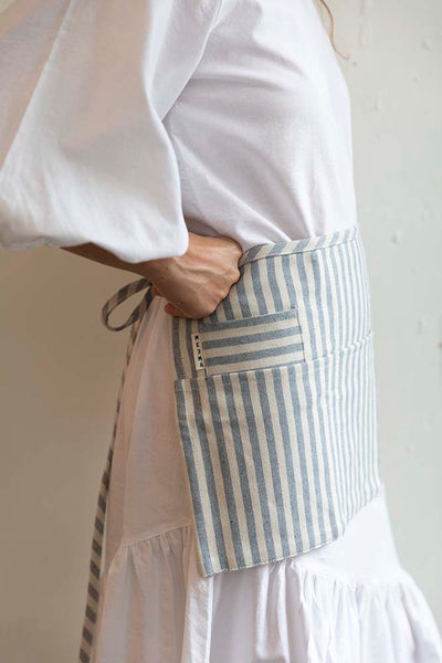 Side view of the Striped Waist Apron in a light blue denim color and white stripes has long straps to tie at your desired fit