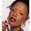 Dark skinned model wears the Genet multi bene stick on her lips and cheeks while putting the lipstick on and looking into the camera. A deep berry color.