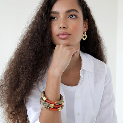 Curly haired brunette model wears a stack of 3 handmade bracelets. The Pichulik Aruba Bracelet in brown and red. Thin rope assembled with brass caps and embellished with wax cord.